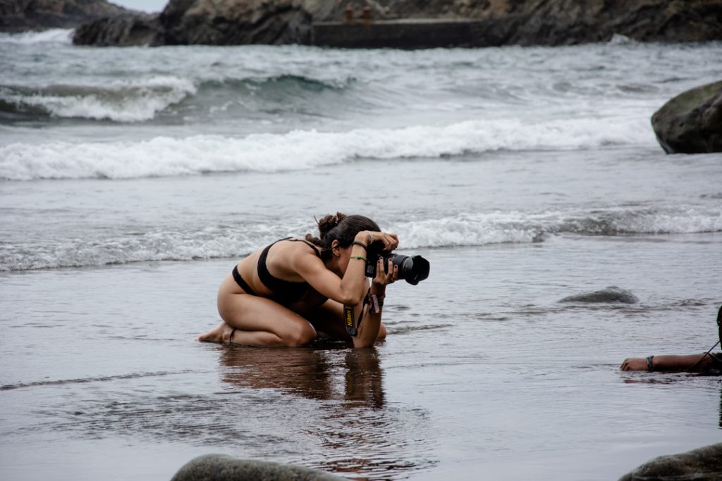 Fotografía en la que vemos a una fotógrafa, haciendo fotos a una modelo en la playa, se ven las olas al fondo, detrás de la fotógrafa. La fotógrafa es Elia Gil