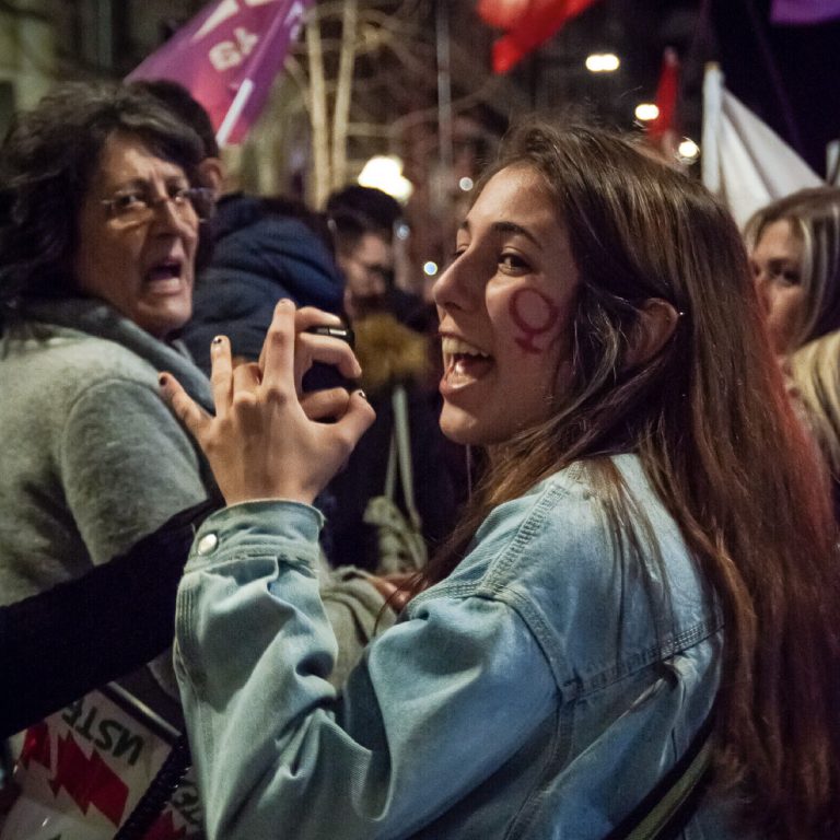 Mujer en manifestación feminista sujetando un megáfono y gritando a través de él. También vemos una mujer mayor al fondo uniéndose al grito.
